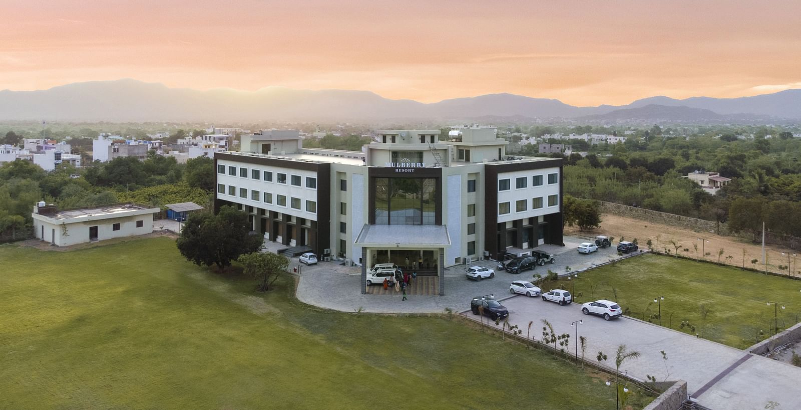 An aerial view of the Bamboo Saa Mulberry Resort, Pushkar at sunset or sunrise surrounded by green lawn and the backdrop of hills.