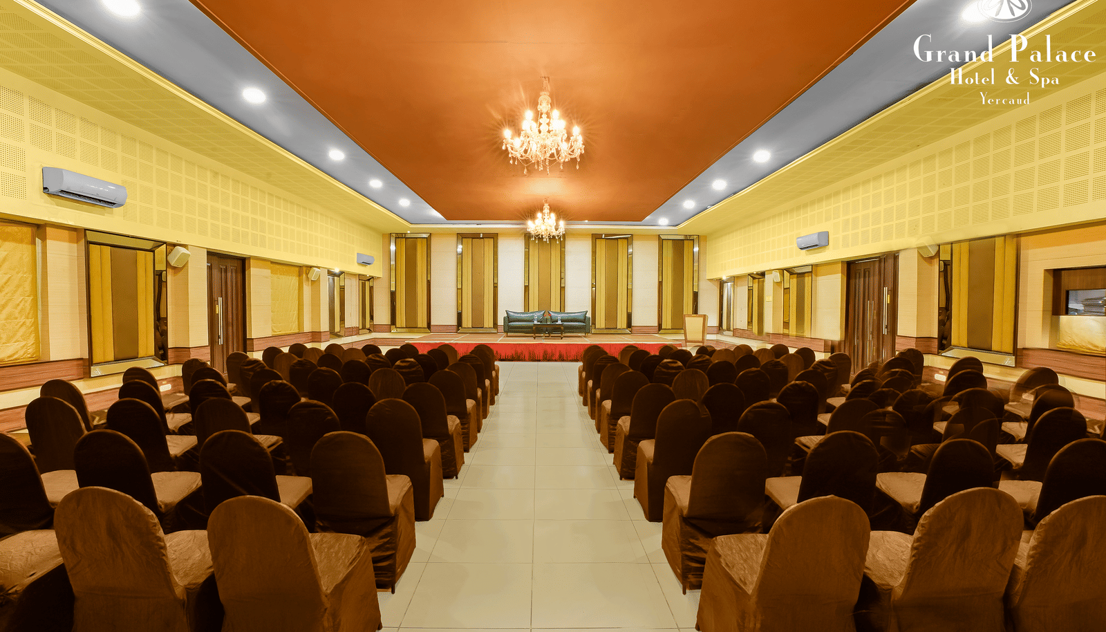 Grand I hall at Grand Palace, Yercaud with rows of chairs, centre aisle, chandeliers, projector screens and high ceiling suitable for meetings or seminars.