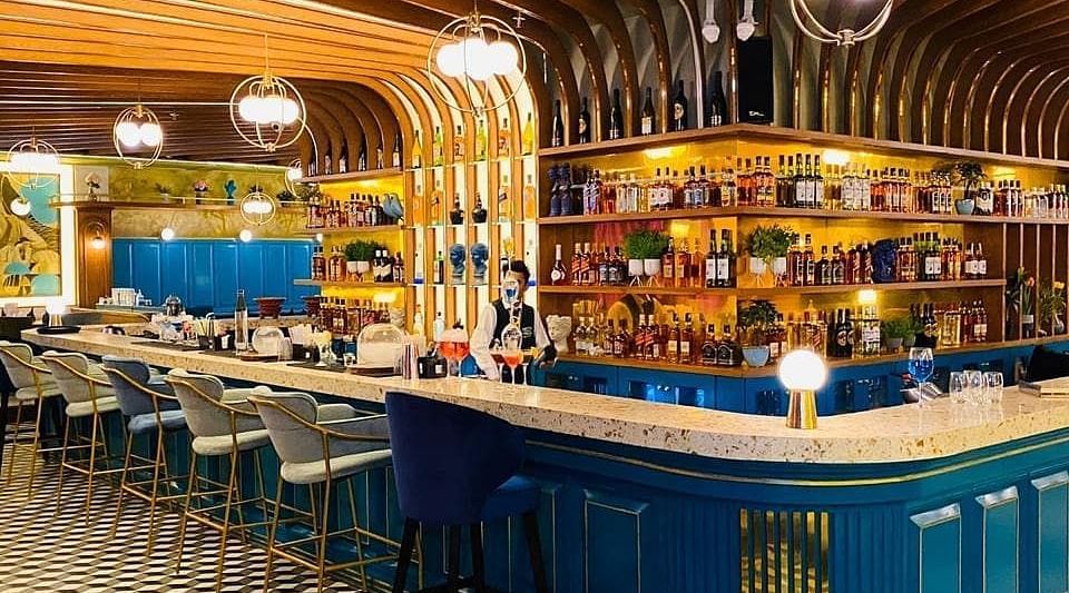An image of a bar featuring a counter table, chairs, and a shelf where liquor items are placed - Themis Barbecue House, Panipat