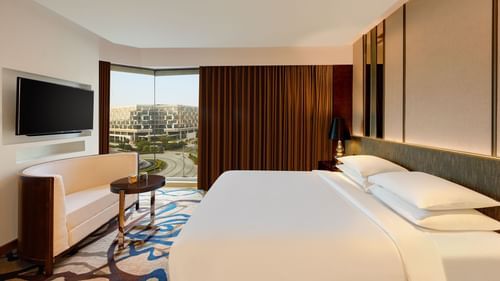 Image of Luxury Suite Room at Pride Plaza Hotel, New Delhi featuring a plush bed in front of a TV mounted on the wall below which a seating space is arranged next to a large window partially covered by curtains