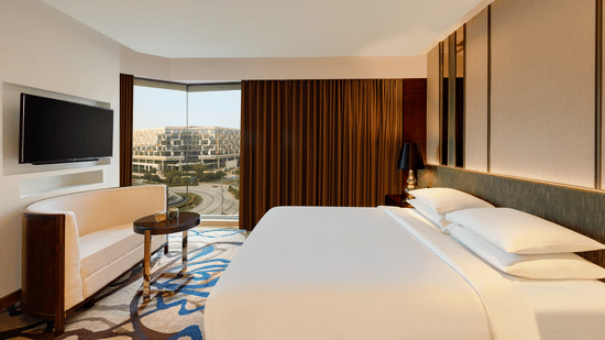 Luxury Suite Room at Pride Plaza Hotel, New Delhi featuring a plush bed in front of a TV mounted on the wall below which a seating space is arranged next to a large window partially covered by curtains