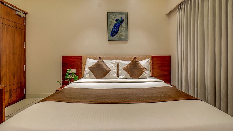 A spacious room at Harigovinda's JNStay with a large bed, wooden headboard, bedside table, curtains pulled to the side, and a photo frame above the headboard