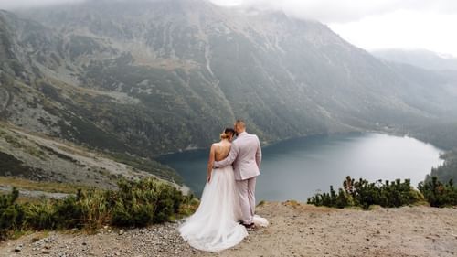 A wedding couple hugging each other on top of a mountain overlooking a lake by the valley.