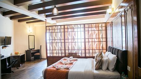 A room at New Orchid Lords Inn Gangtok contains a bed with pillows and towel swans, a bedside table with water bottles, a desk with a mirror and chair, and a TV.