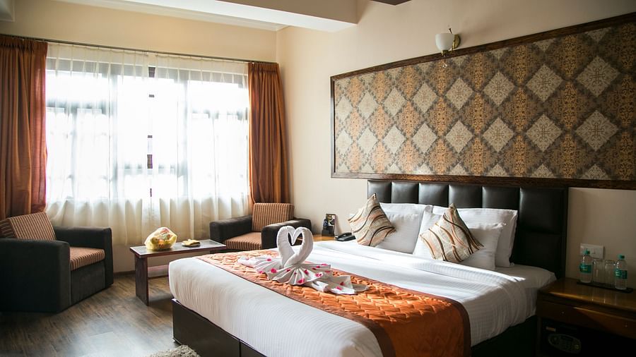 A room at New Orchid Lords Inn Gangtok contains a bed with pillows and towel swans, bedside tables with lamps and water, chairs, a coffee table with flowers, curtains, and a rug.