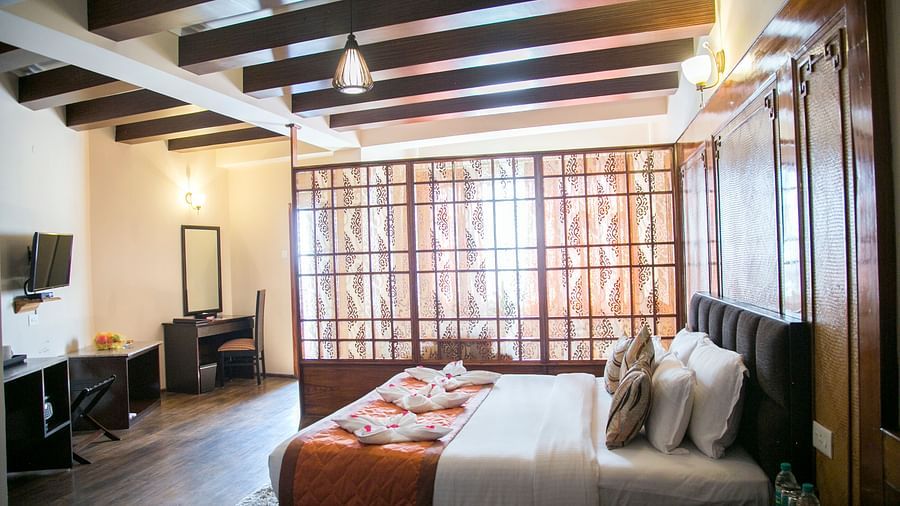 A room at New Orchid Lords Inn Gangtok contains a bed with pillows and towel swans, a bedside table with water bottles, a desk with a mirror and chair, and a TV.