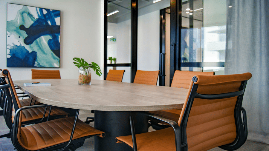 Modern conference room with a round wooden table, brown leather chairs, glass partition walls, and a small indoor plant in a contemporary office setting.