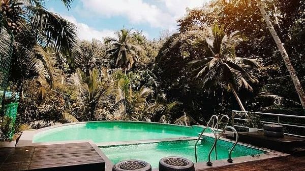 An outdoor swimming pool surrounded by tropical palm trees, with a wooden deck in the foreground featuring some small decorative items - Sea Hills Hotels, Sri Vijaya Puram (Port Blair)