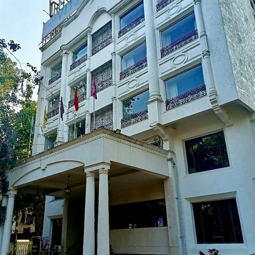  Exterior view of a multi-storey white building with large glass windows and a covered entrance supported by columns, surrounded by trees.