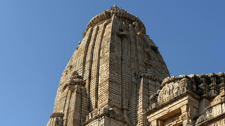 A soaring, intricately carved stone temple tower stands under a bright blue sky, captured from a low angle.