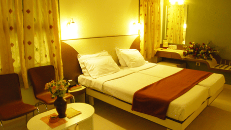 A comfortable hotel room with a large double bed, white linens, warm yellow lighting, and a small coffee table with chairs - Hotel Pandian