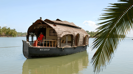 Traditional houseboat with a thatched roof floating on the water, partially framed by palm leaves at Paradise Lagoon Resort, Udupi.