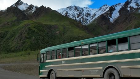 Green bus travelling through a road with green mountains in the background and snow on peak of the mountains.