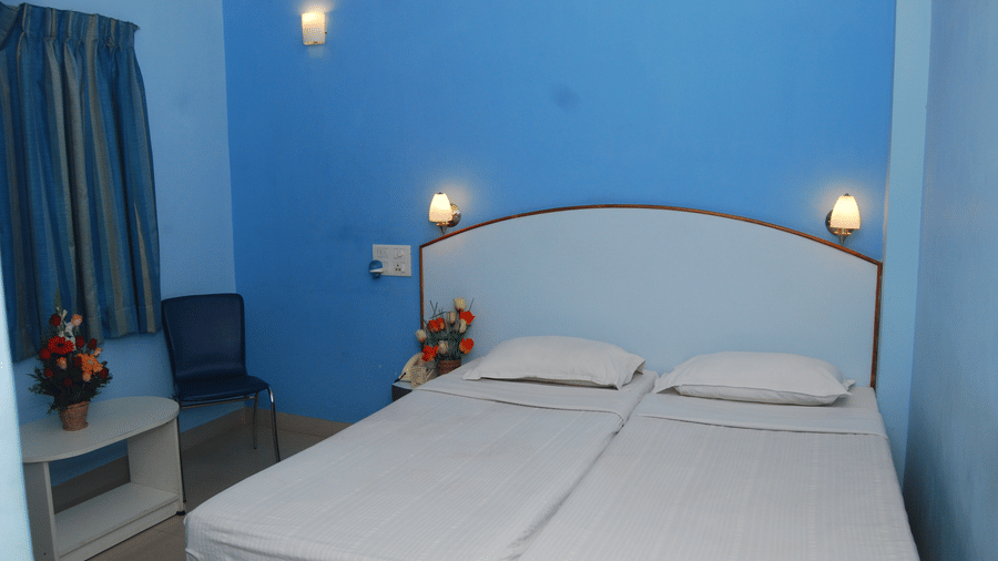 A clean hotel room setup featuring 2 single beds placed side by side against a bright blue wall with bedside lamps - Hotel Pandian