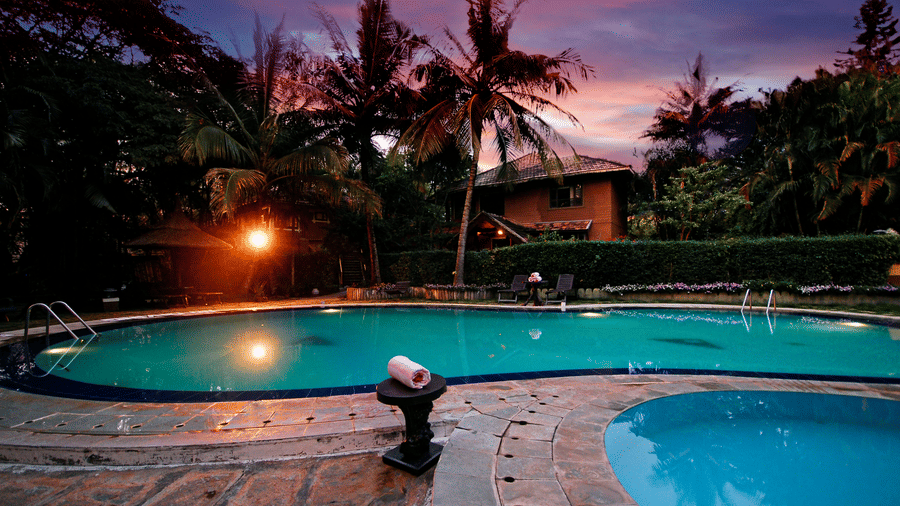 Outdoor swimming pool at Hoysala Village Resort, Hassan reflecting a warm sunset, surrounded by palm trees and a low-rise building at dusk.