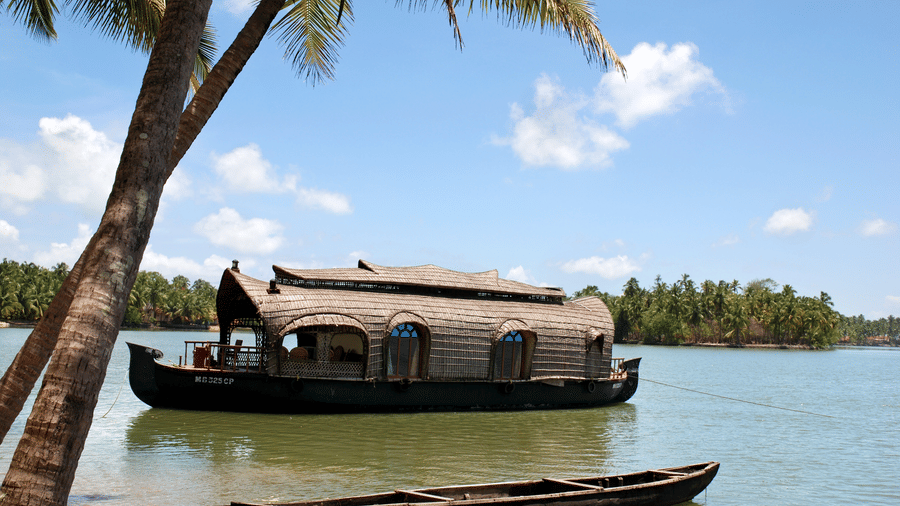 Docked houseboat with a curved roof on calm backwaters, with trees and sky in the background at Paradise Lagoon Resort.