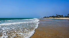 A view of Varca Beach, one of the most popular beaches in South Goa, with the foamy waves crashing on the shoreline with trees in the distance and blue sky above.