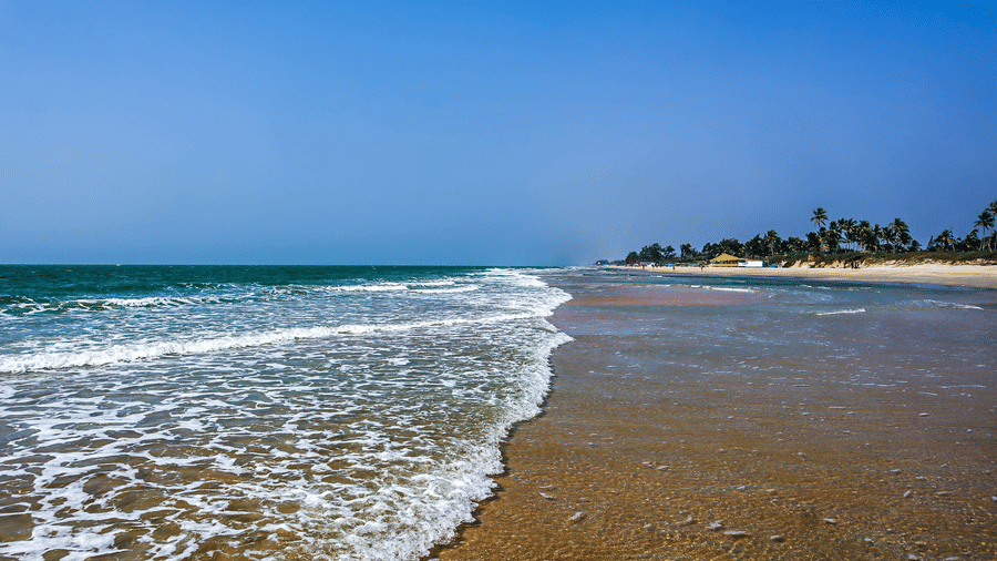 A view of Varca Beach, one of the most popular beaches in South Goa, with the foamy waves crashing on the shoreline with trees in the distance and blue sky above.