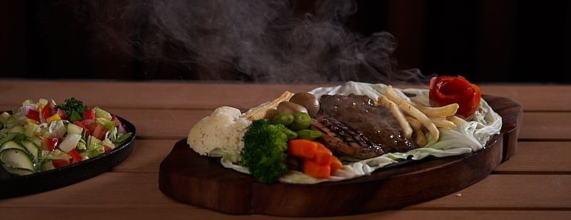  A sizzling hot plate of grilled meat and vegetables with steam rising, served on a wooden platter, accompanied by a side of fresh salad at The Hamuse Hotel, Kodaikanal.