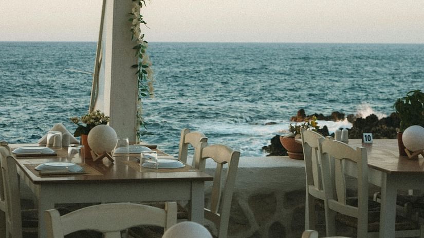 A serene seaside restaurant with wooden tables and chairs, softly lit lamps, and a beautiful ocean view under a pastel sky.