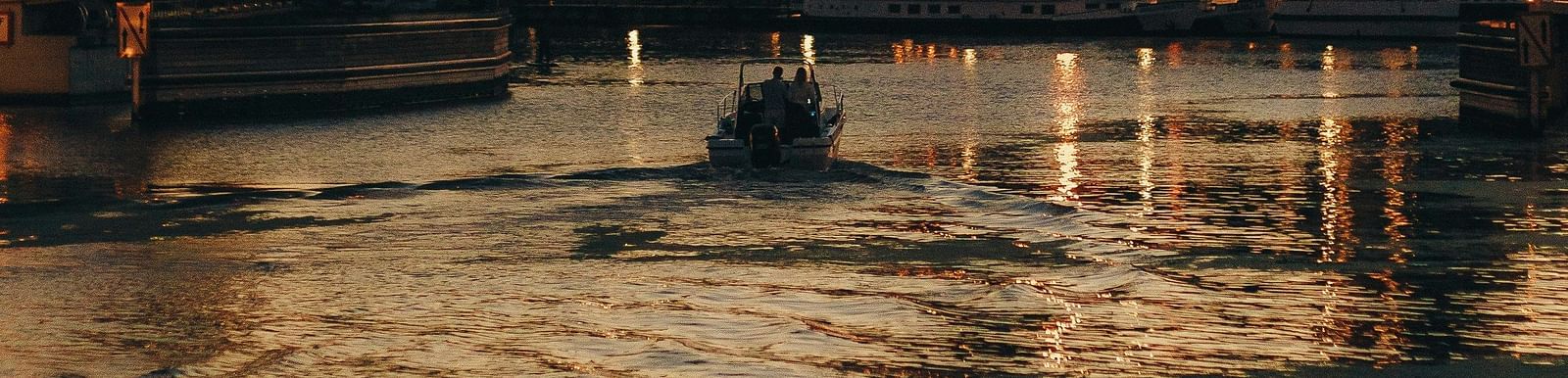 A scenic riverside at sunset with a raised bridge, historic buildings, and a boat gliding through the golden-lit waters.