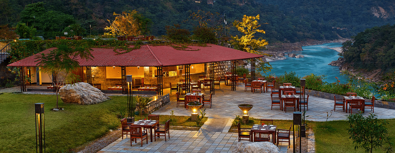 An image of a well lit restaurant surrounded by lush greenery with a view of a river - Leisure Hotels