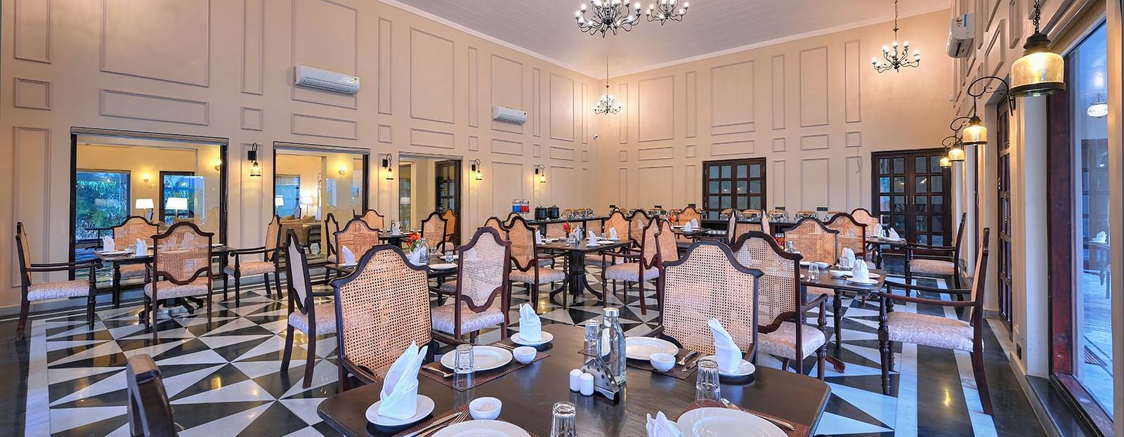 Elegant dining hall with chandeliers, classic decor, and neatly arranged tables set with fine dining essentials. Large windows and warm lighting create an inviting ambience - Singhvilas, Ranthambore