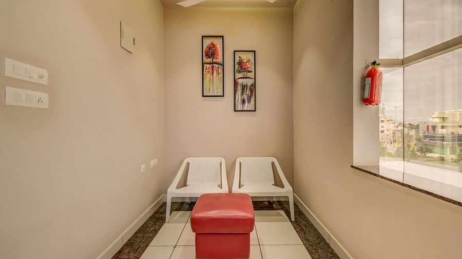 Two chairs placed beside a small cushioned table near a large glass window, with wall-mounted photo frames above, at Harigovinda's JNStay