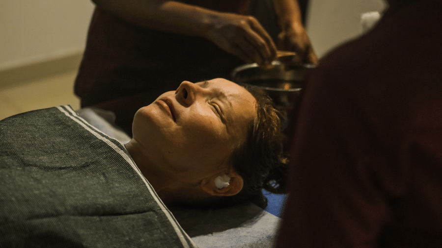 A woman lying down on the massage bed with two masseurs next to her at Ayur On The Beach Nattika. 
