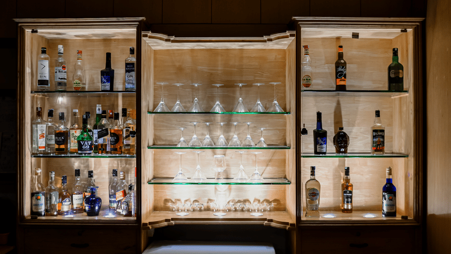 Illuminated wooden display cabinet with glass shelves holding liquor bottles and wine glasses at Kadkani River Resort, Coorg.