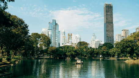 A scenic view of a city park with a calm lake in the foreground, lush green trees lining the water, and modern high-rise buildings, including a prominent thin white tower, in the background under a blue sky.