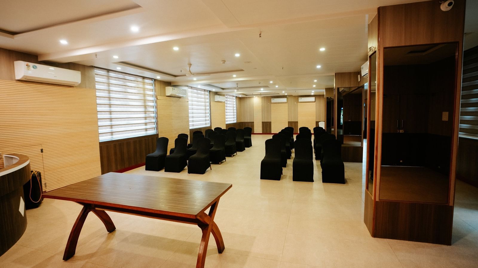 Panoramic view showing the dark wood table and the far end of the hall with built-in wooden shelving or cabinetry at Daksh The Madhuvan Suites, Dwarka.