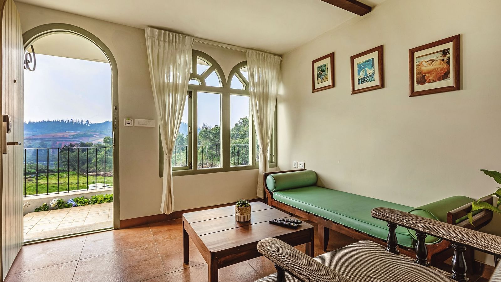 Living room at Mango Hill Shola, Ooty, with cushioned seating, wooden furniture, and arched balcony windows offering panoramic views of the scenic outdoors.