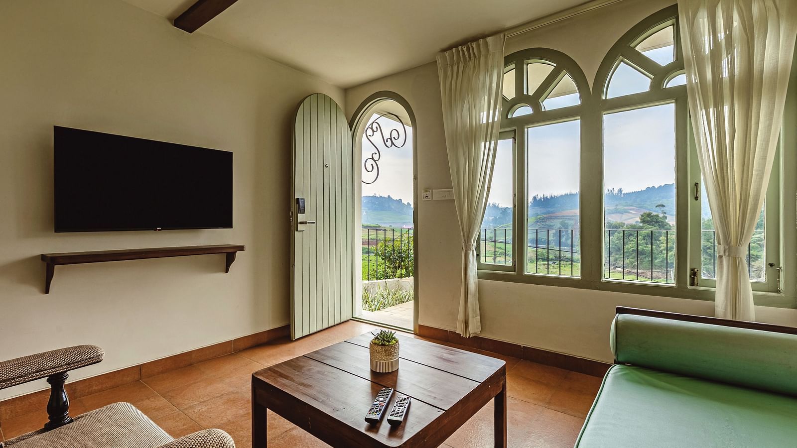 Inviting living area at Mango Hill Shola, Ooty, featuring a mounted television, cushioned seating, and large arched windows letting in ample natural light.