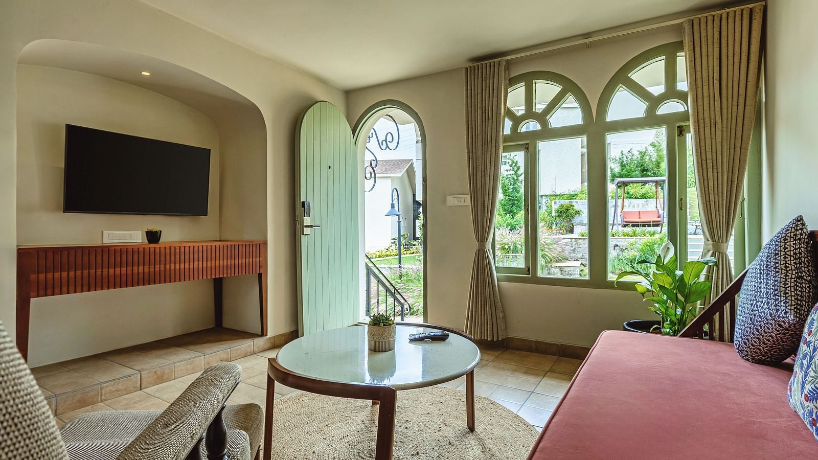 Spacious living room with a red sofa, TV, and arched windows overlooking the garden at Mango Hill Shola, Ooty.