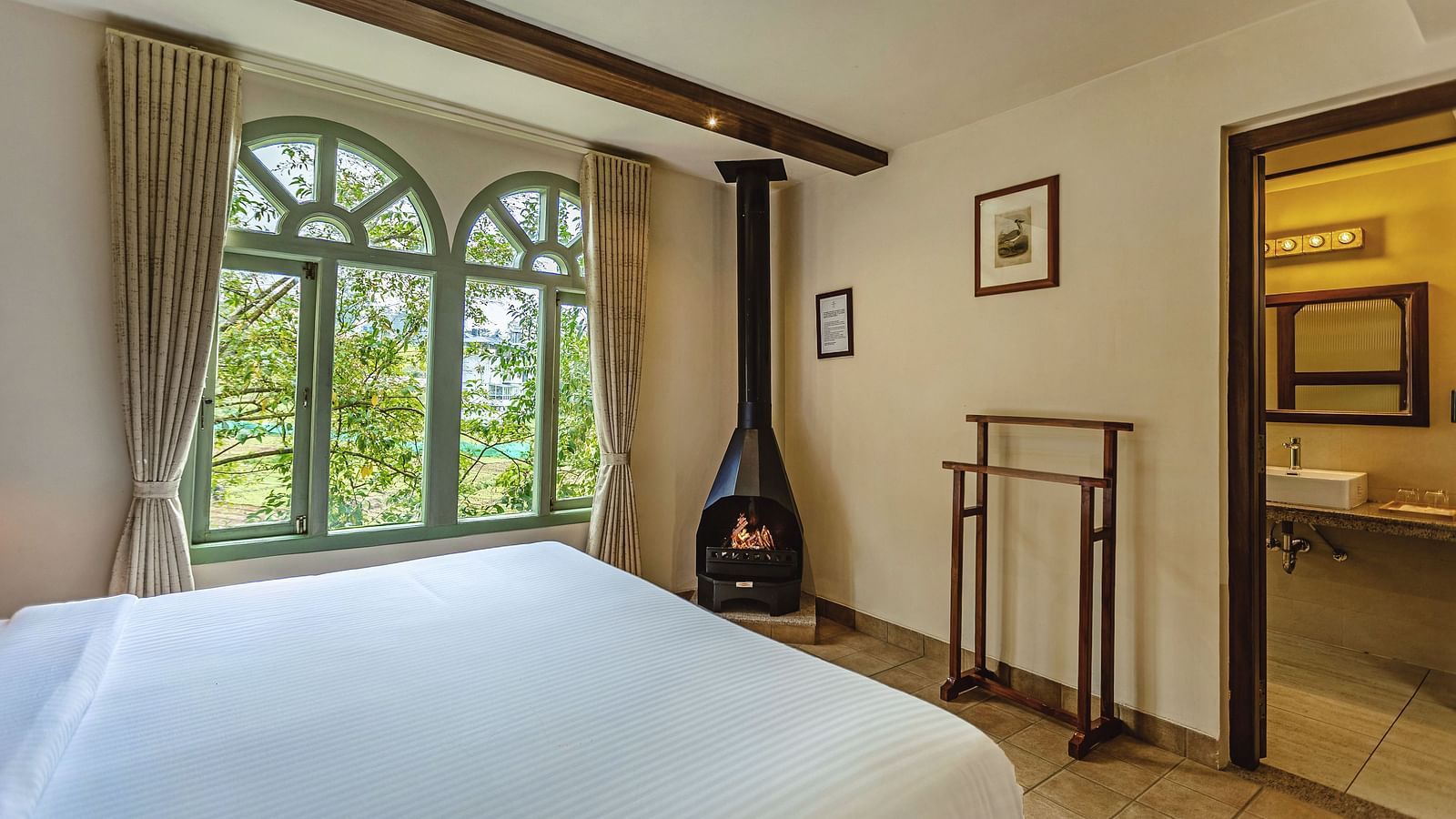  A corner view of a warm and elegant bedroom at Mango Hill Shola, Ooty with a central fireplace and peaceful outdoor scenery visible through the windows.