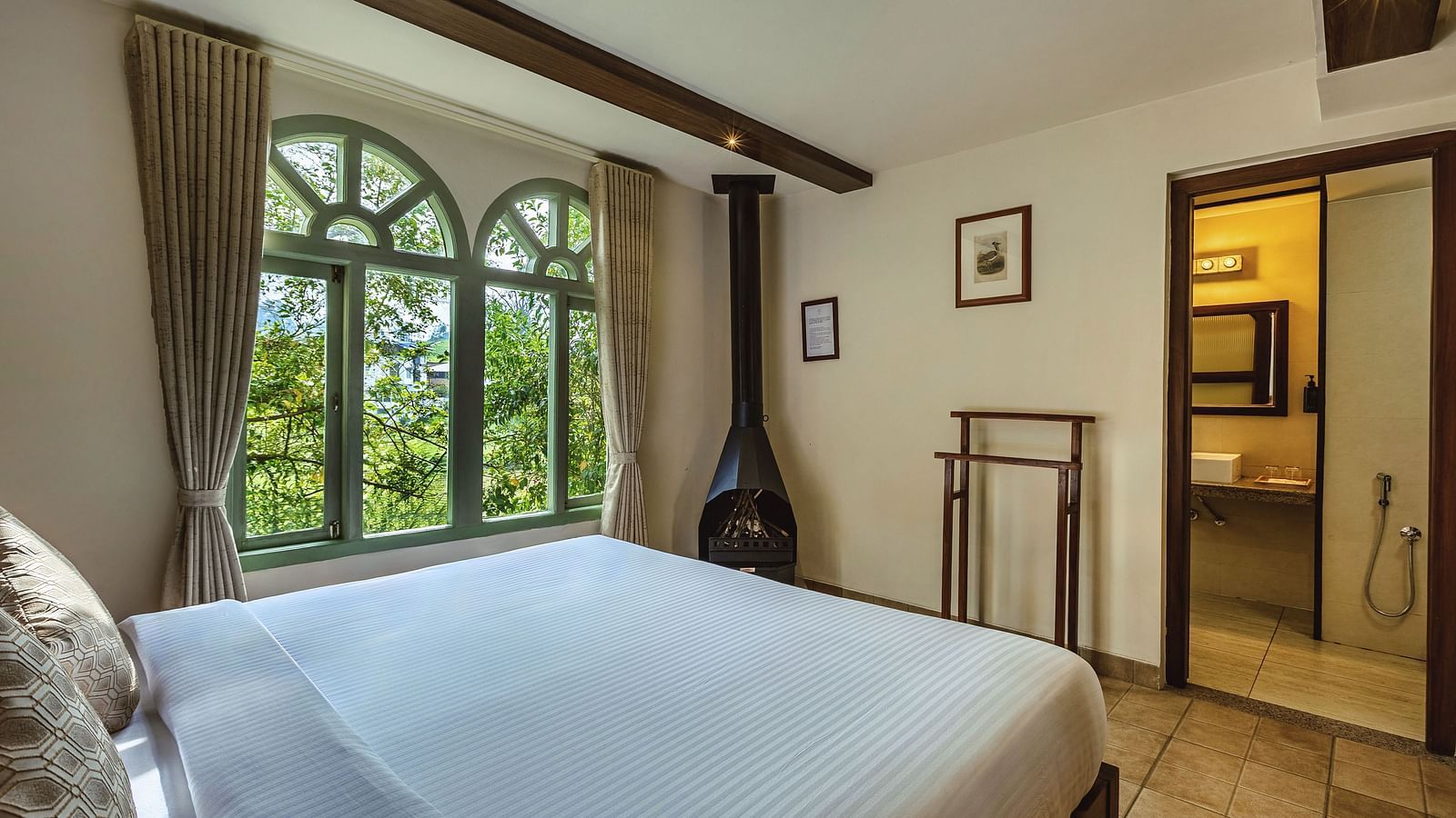 Well-lit bedroom with a queen bed, classic furniture, and garden views through a large arched window at Mango Hill Shola, Ooty.