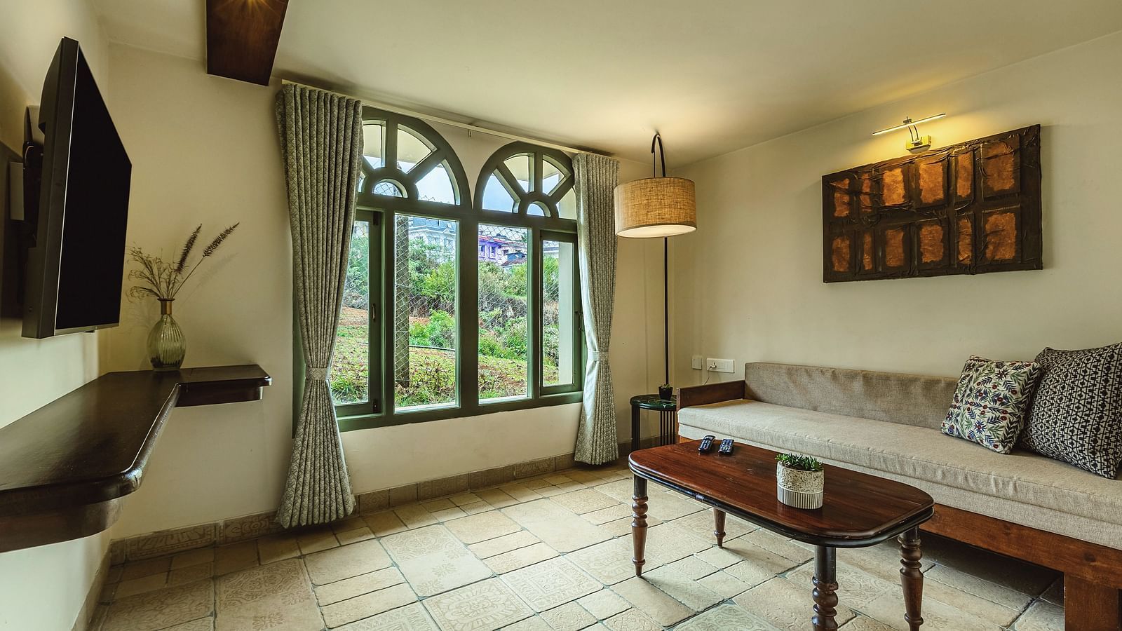 Spacious studio room's seating area at Mango Hill Shola, Ooty with large arched windows, a wooden daybed, wall-mounted TV, and serene plant and trees along with that buildings.