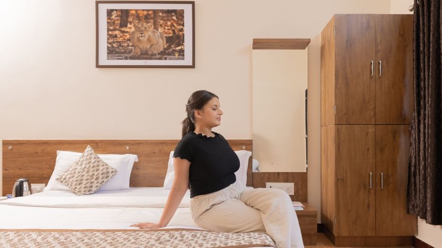 A woman sitting on a bed with a wall art above it featuring a mirror and a wooden wardrobe at Daksh Resort & Amusement Park, Sasan Gir.