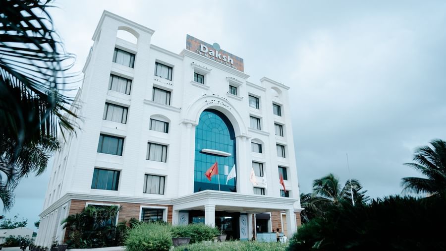 Large, modern white hotel building exterior on a slightly overcast day. The main entrance features a large arched blue glass window at Daksh The Madhuvan Suites, Dwarka.