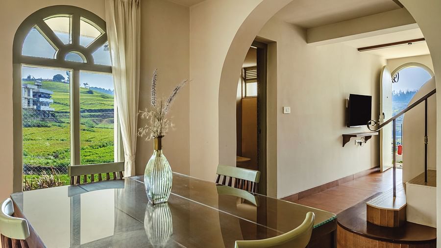 dining space at Mango Hill Shola, Ooty, featuring elegant furniture and a large arched window that frames a  hillside & building view.