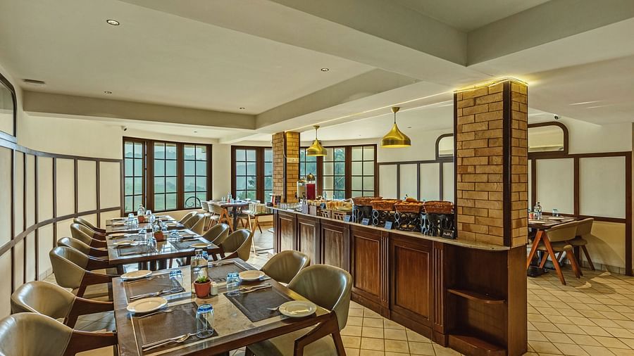 An indoor dining area at Mango Hill Shola, Ooty with glass-topped tables, cushioned chairs, and large windows bringing in natural light.