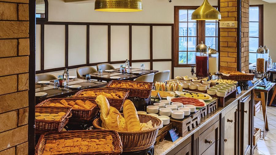 Buffet counter at Mango Hill Shola, Ooty showcasing a wide variety of fresh breads and breakfast items, set under warm pendant lighting.