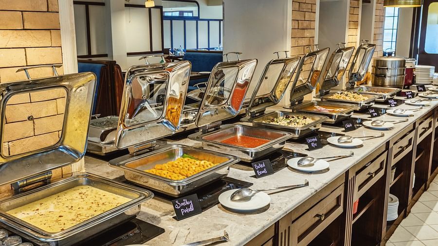 Buffet line with stainless steel chafing dishes offering a variety of hot meals at Mango Hill Shola, Ooty, set against brick and wood interiors.