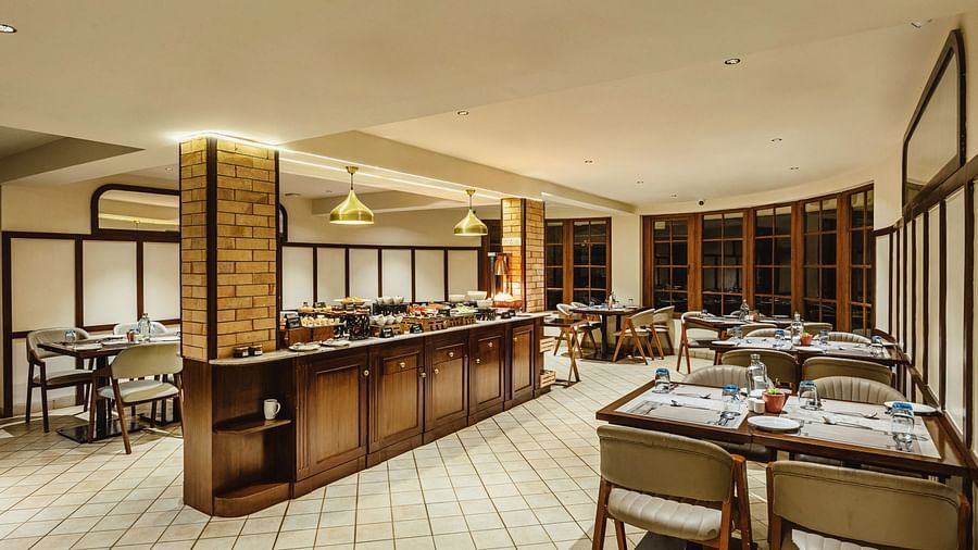 Warm and inviting dining setup at Mango Hill Shola, Ooty, featuring neatly arranged tables with cutlery, wooden cabinetry, and a central buffet counter illuminated by pendant lighting.