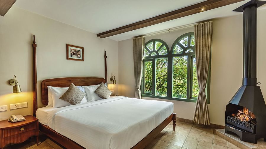 Sunlit bedroom at Mango Hill Shola, Ooty with a wooden bed, side tables, and large arched windows offering lush green views.