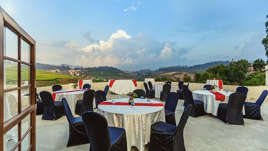 Rooftop event setup at Mango Hill Shola, Ooty, featuring round tables covered in white linens with red and black accents, surrounded by black banquet chairs, overlooking lush green hills and a dramatic cloudy sky.