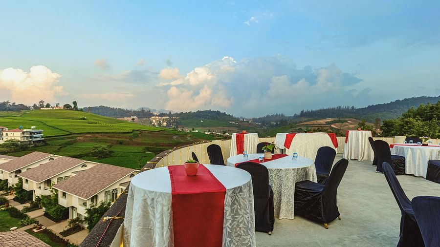Scenic rooftop dining and event area at Mango Hill Shola, Ooty, showcasing neatly arranged tables with red and white covers, black chairs, and panoramic views of rolling hills, terraced fields, and a clear sky.