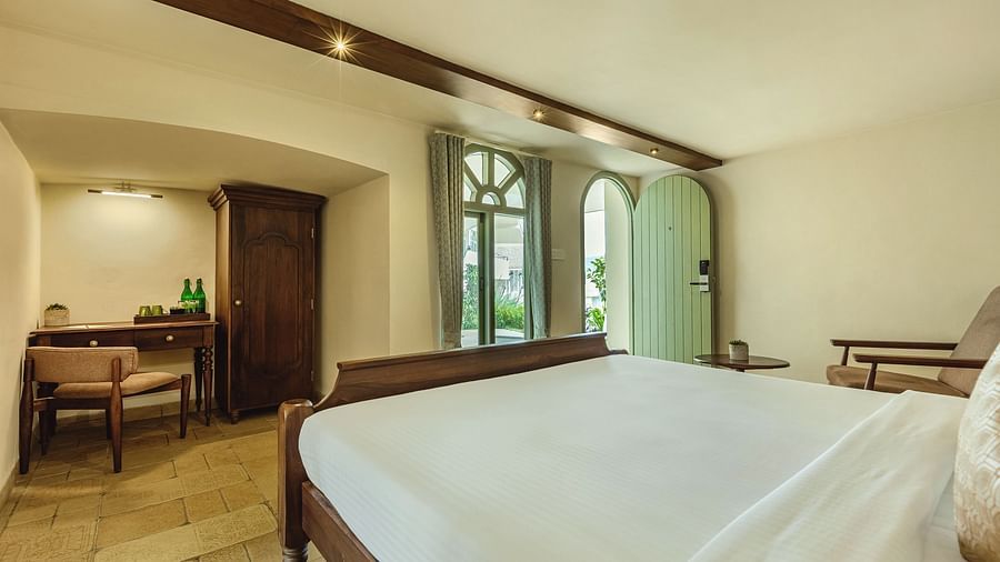 Studio room at Mango Hill Shola, Ooty showcasing a classic wooden bed, arched window, writing desk, and a serene view of the outdoors.