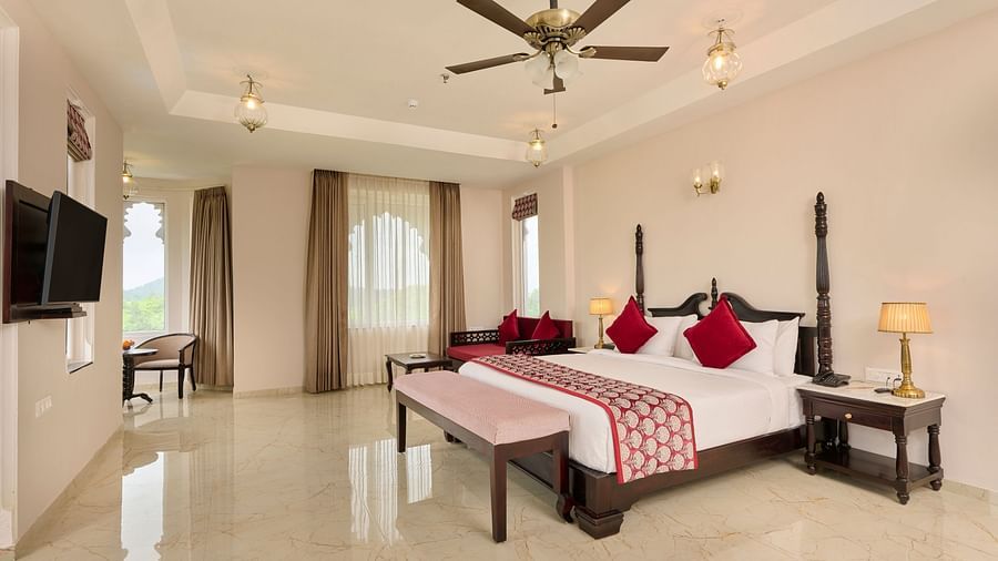 Luxurious hotel room with a four-poster bed, red accents, a seating area, and a flat-screen TV, bathed in natural light.